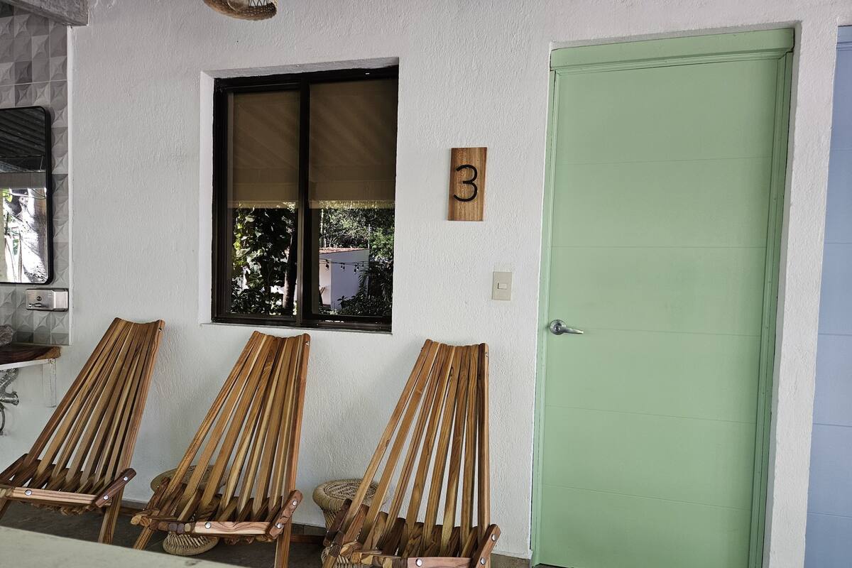 A room with three wooden chairs, a green door, and a window with a view of greenery.