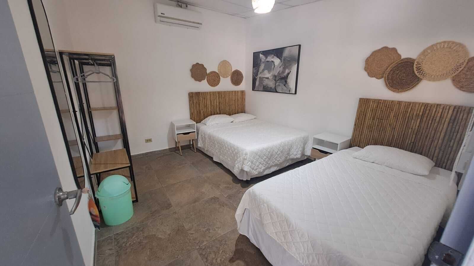 Quarto com duas camas, uma mesinha, ar-condicionado fixado na parede e uma porta com porta-toalhas.