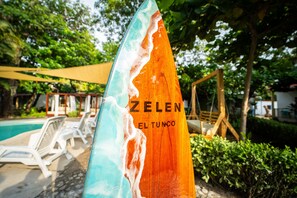 Une planche de surf en bois sur laquelle on peut lire « ZELEN EL TUNCO », le tout dans un décor de piscine avec des chaises longues et de la verdure.