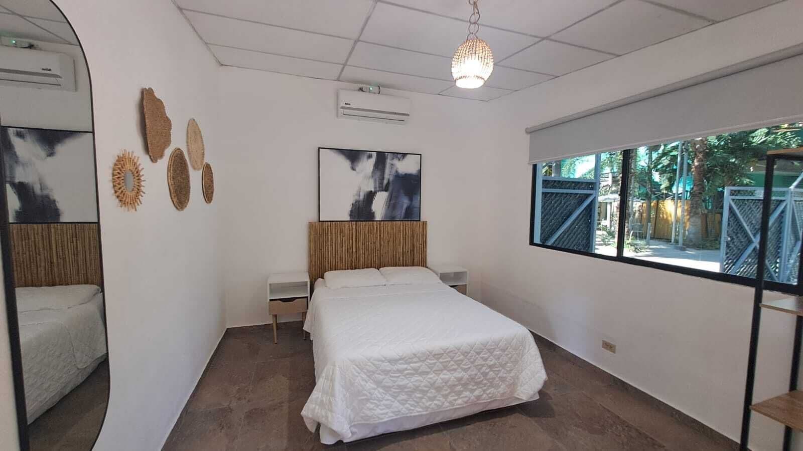 Un dormitorio con cama, mesita de noche, aire acondicionado instalado en la pared, ventana con vistas a la vegetación y decoración en las paredes.