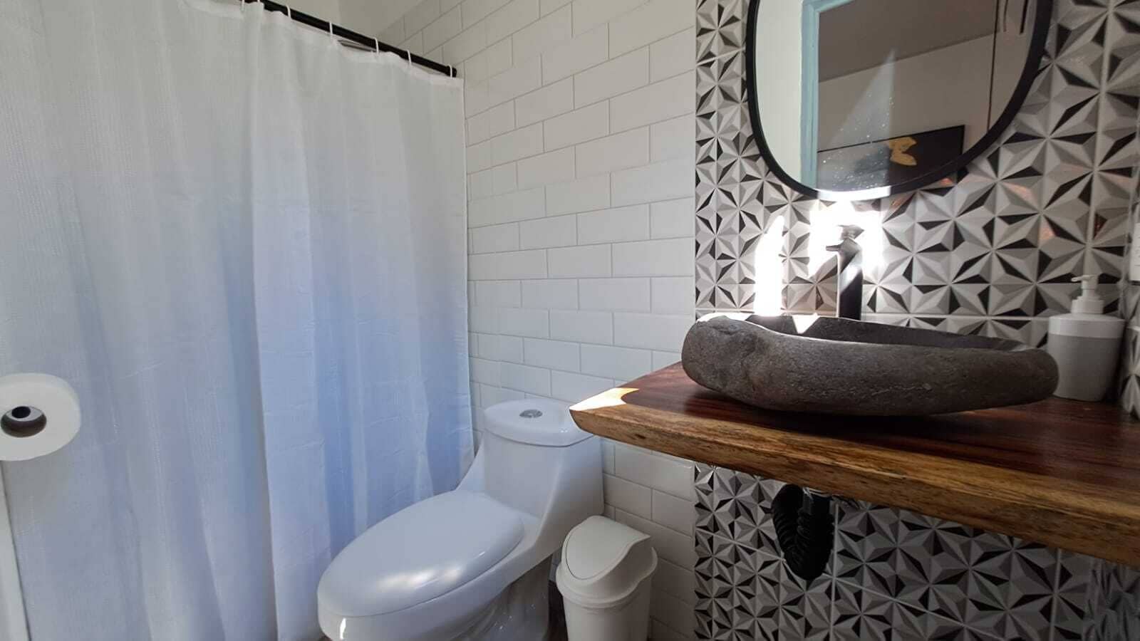 A bathroom with a white toilet, a shower curtain, a sink with a stone basin, and a mirror with a geometric tile backsplash.