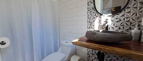 A bathroom with a white toilet, a shower curtain, a sink with a stone basin, and a mirror with a geometric tile backsplash.
