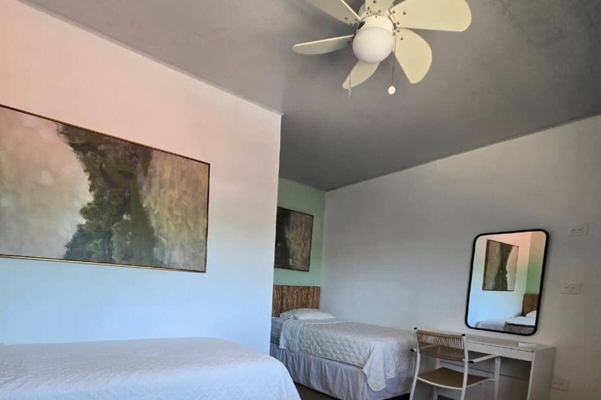 A bedroom with two beds, a desk, a chair, a ceiling fan, and a painting on the wall.