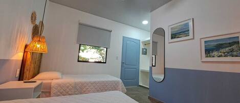 A bedroom with two beds, a bedside table, a lamp, a window, a door, and wall-mounted artwork.