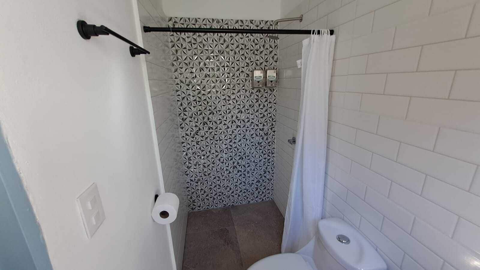 A small bathroom with a shower, white tiled walls, a toilet, and a roll of toilet paper on the wall.