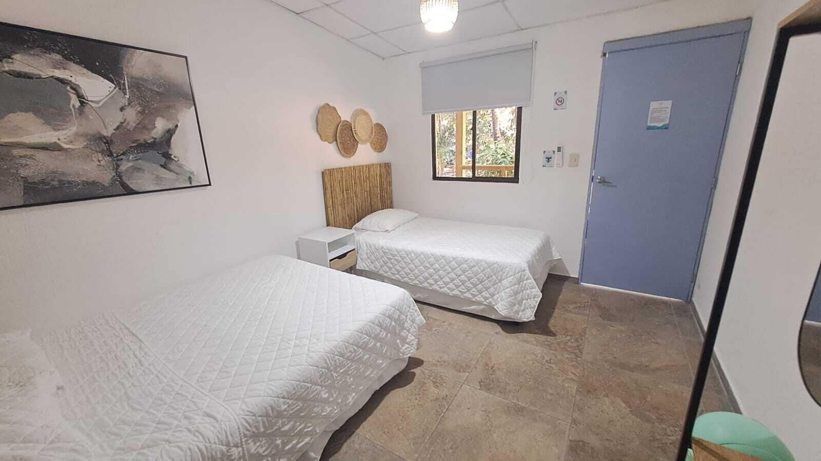 Una habitación con dos camas, una ventana con persianas, una puerta y una obra de arte colgada en la pared.