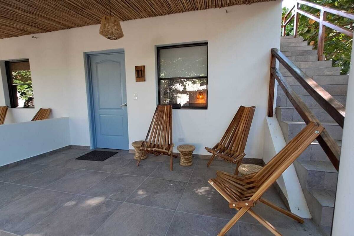A covered outdoor area with a blue door, wooden chairs, a window, and a staircase.