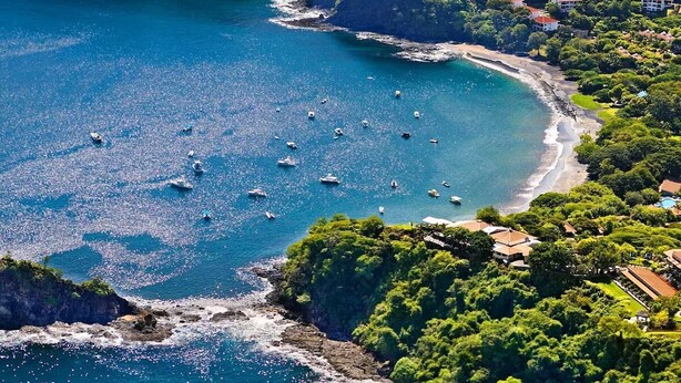 An aerial view of a coastal area with numerous boats on the water, a sandy beach, and lush greenery.