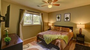 Un dormitorio con cama, lámparas de noche, ventilador de techo, ventana con cortinas y una pequeña mesa con una planta.
