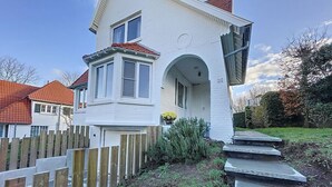 Ein weißes Haus mit rotem Dach, Holzzaun und einer Betontreppe, die zur Haustür führt.