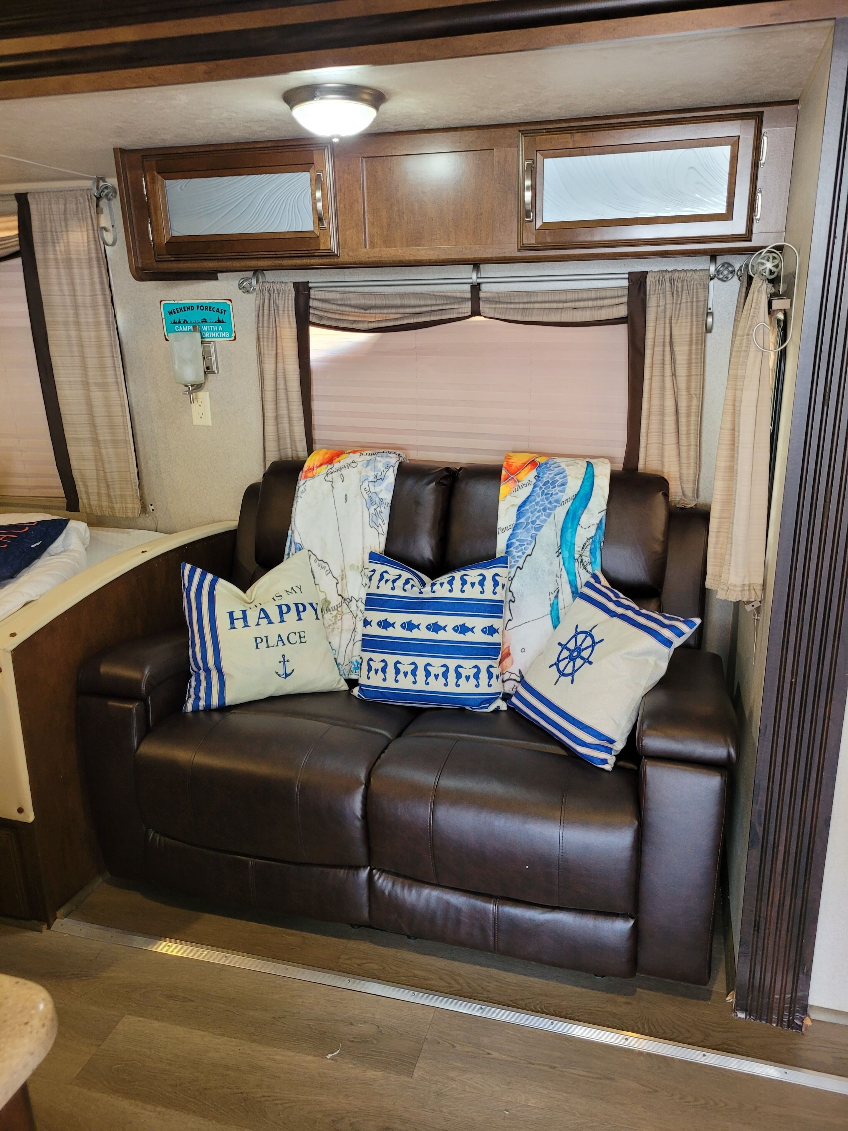 A cozy RV interior with a brown leather sofa, a mounted TV, and a window with curtains.