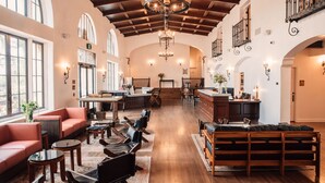 Een ruime lobby met een hoge plafond, houten balken en een centrale kroonluchter. Er zijn zitgedeeltes met banken en tafels, en een receptiebalie op de achtergrond.