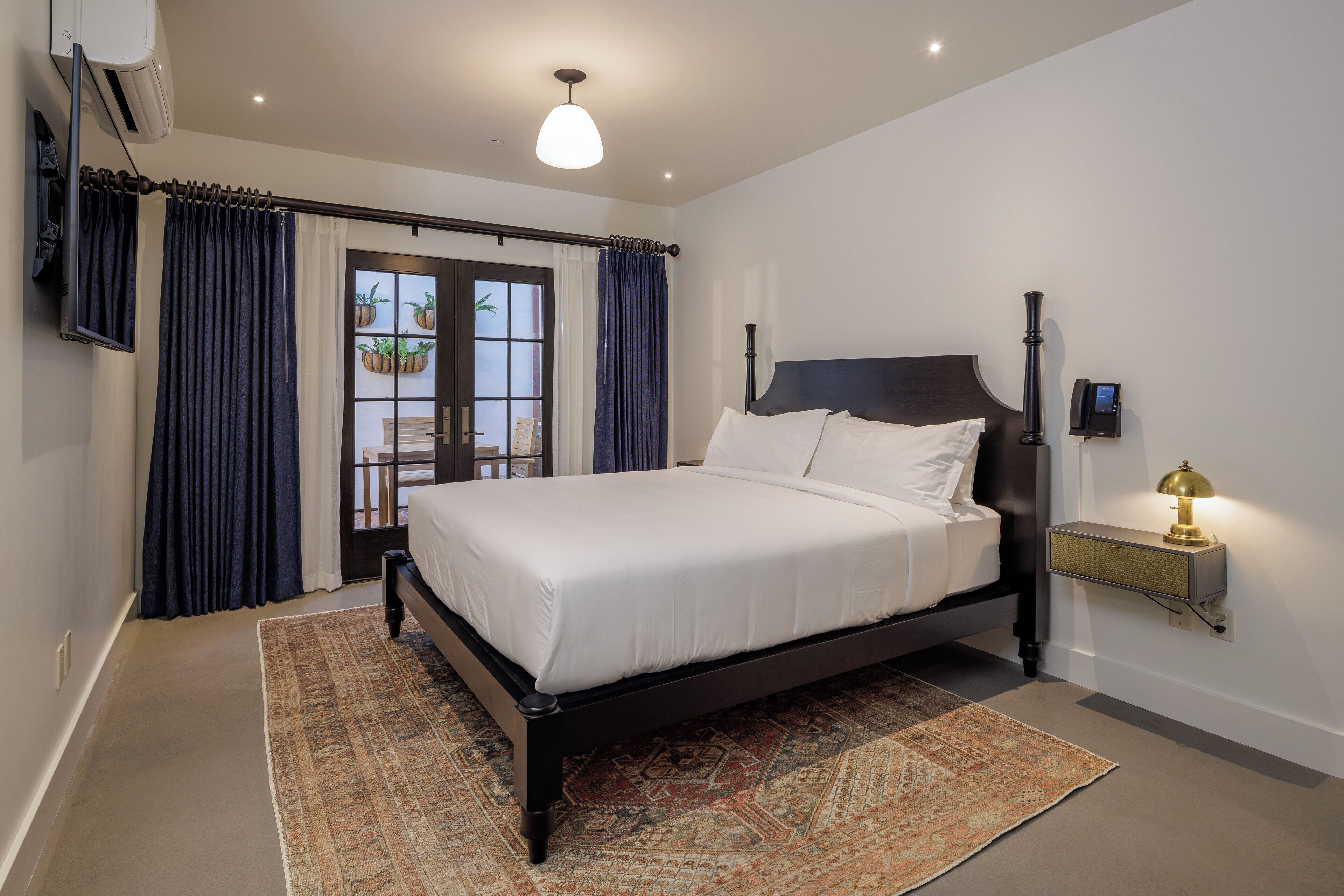 A bedroom with a bed, a nightstand with a lamp, a window with curtains, and a wall-mounted air conditioner.