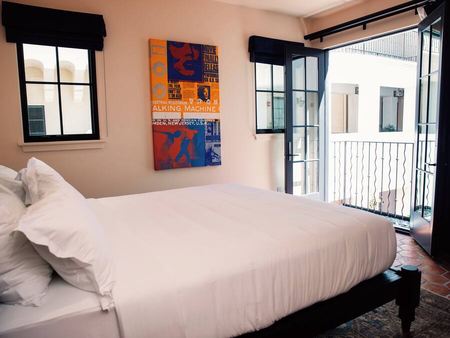 A neatly made bed with white linens, a large window with a black valance, and a colorful poster on the wall.