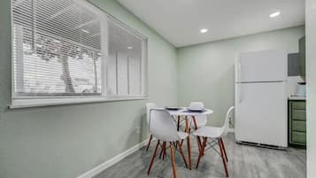 A small kitchen with a white refrigerator, a dining table with four chairs, and a window with blinds.