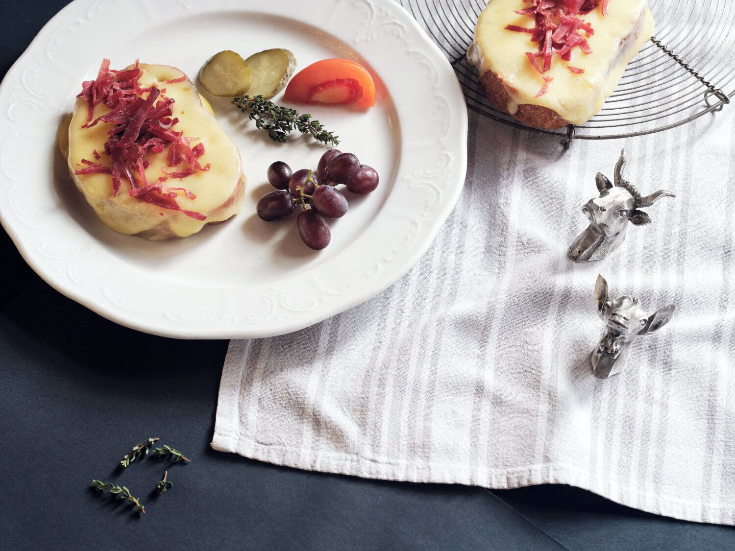 A plate with two open-faced sandwiches topped with cheese and red onions, accompanied by pickles, a slice of tomato, and grapes.