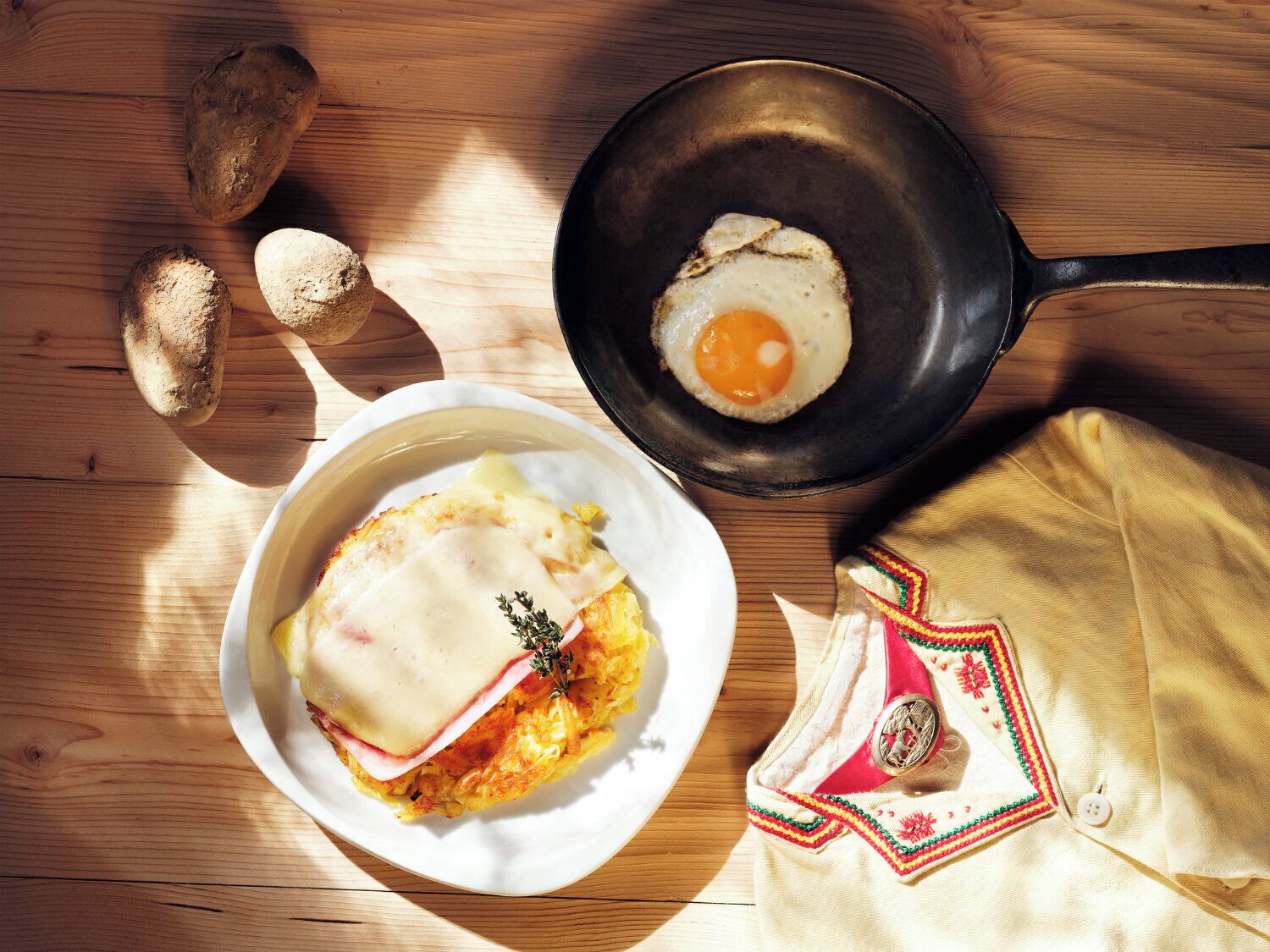 A breakfast plate with eggs, ham, and cheese, a frying pan with an egg, and potatoes on a wooden table.