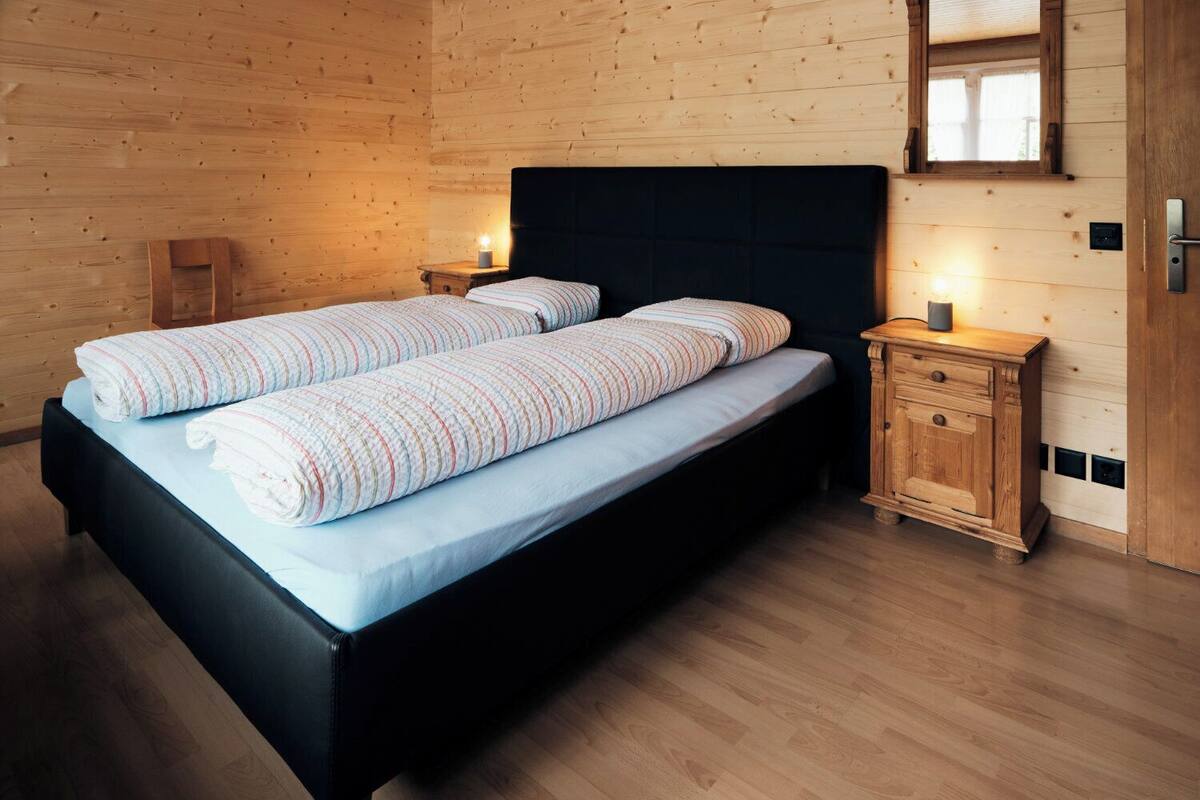 Una cama doble con ropa de cama a rayas, una mesita de noche de madera con lámpara, y un armario de madera en una habitación con paredes y suelos de madera.