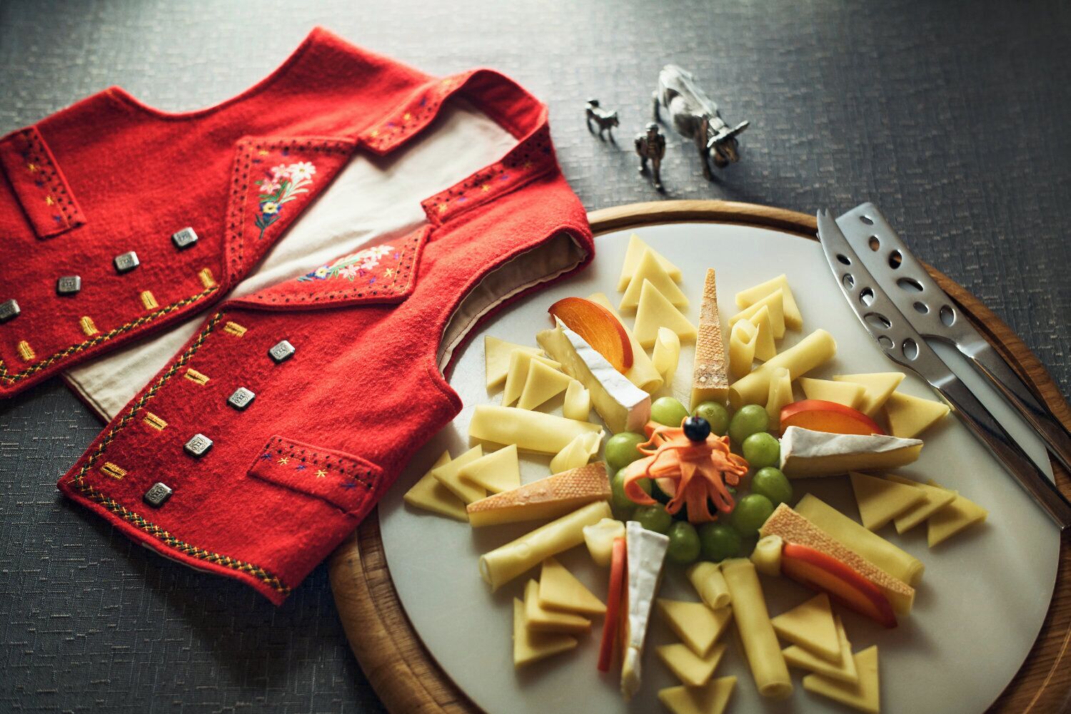 A plate of assorted cheeses and fruits on a wooden board, accompanied by a red garment with decorative stitching.
