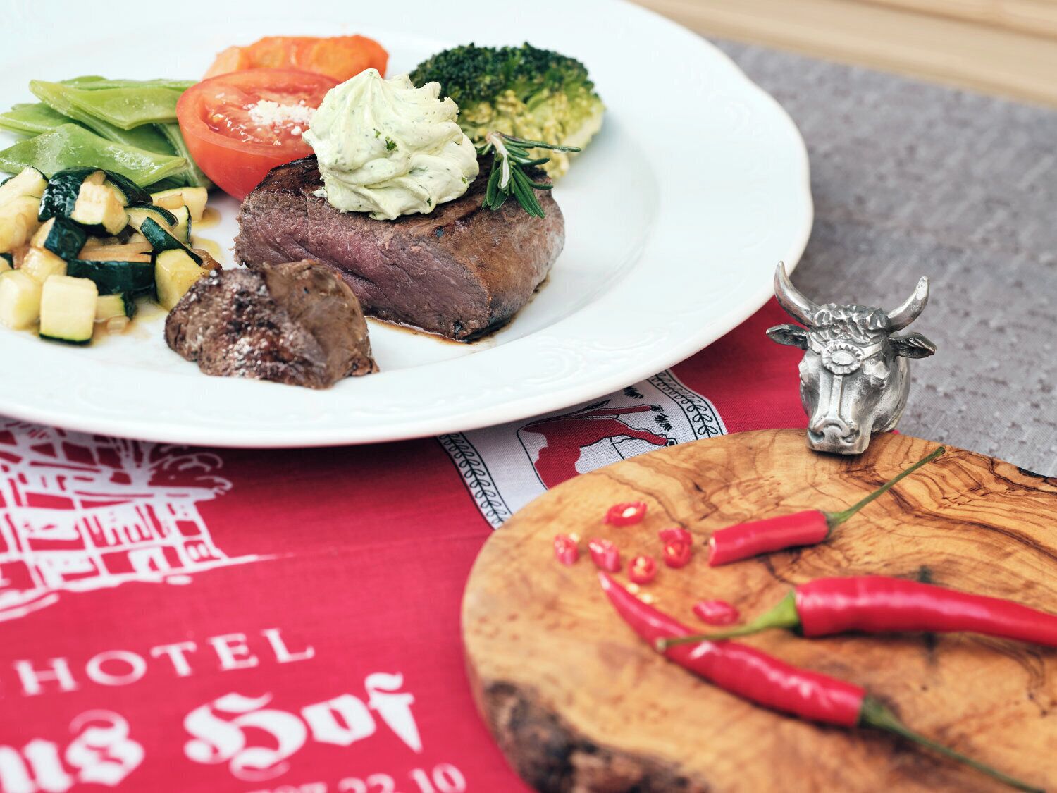 Un plato con bistec, verduras y una cucharada de salsa, una figurita de toro y chiles sobre una tabla de madera.