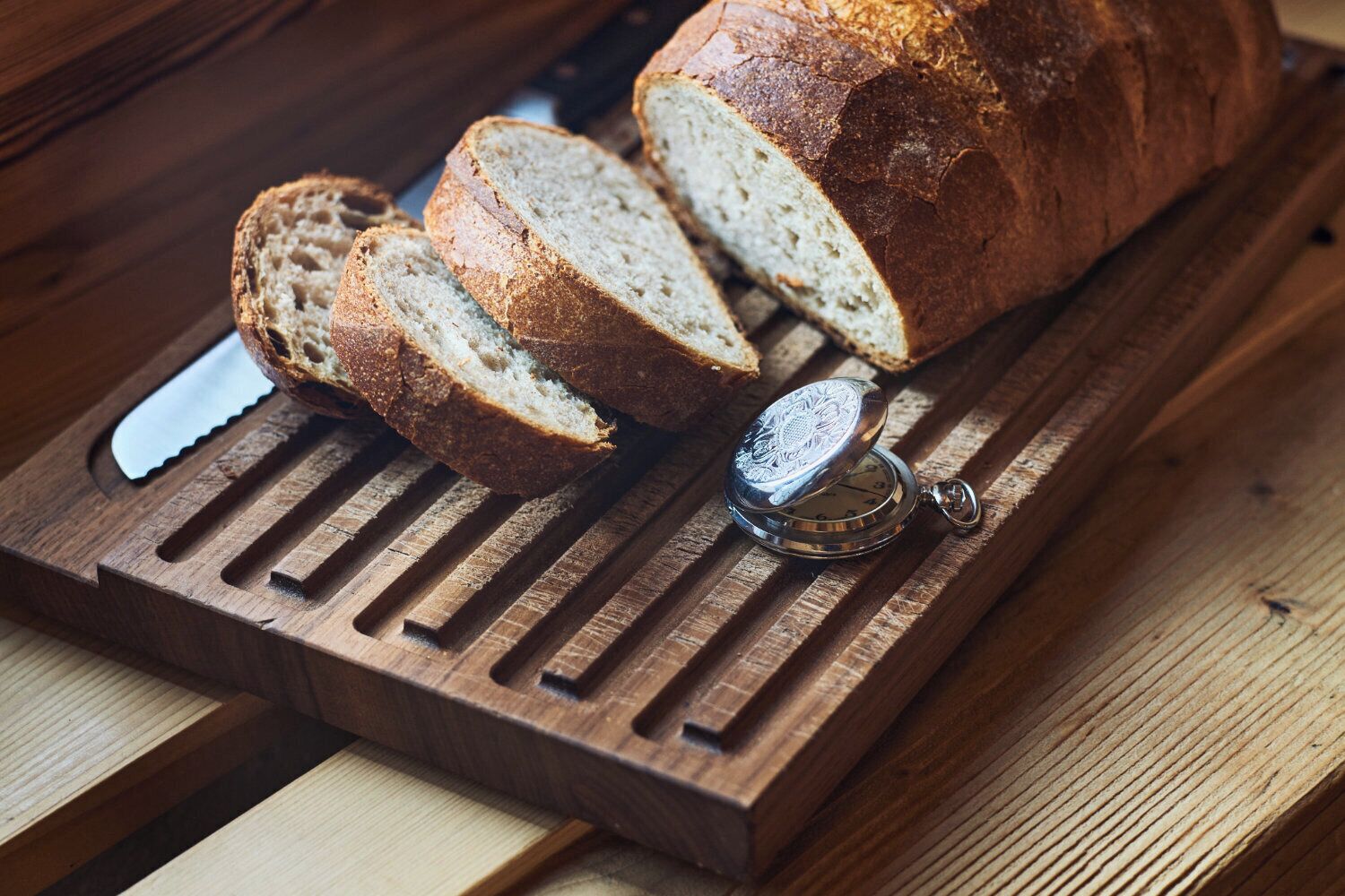 A loaf of bread on a wooden cutting board with slices cut and a pocket watch on top.