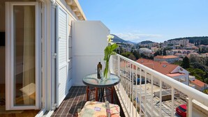A balcony with a table, a vase with flowers, and a bottle. The balcony overlooks a cityscape with buildings and a mountain in the distance. - KABLAR'S SUNNY SUITES 1BR APT with balcony and sea view (Dubrovnik)