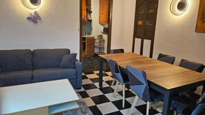 Una habitació amb un sòl a quadradets, una taula de menjador de fusta amb cadires, un sofà i una cuina visible a través d'una porta oberta.