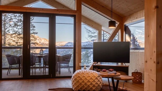 Interior acogedor de una cabaña con piso de madera, un ventanal grande, un televisor y un comedor con sillas y una mesa.