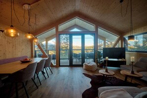 A spacious loft with a high ceiling, a dining area with a wooden table and chairs, a cozy seating area with a sofa and a TV, and large windows offering a view of the outdoors. - Luxury Chalet with Sauna & Hot tub (Steiermark)