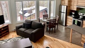 Una sala de estar moderna con un sofá, una mesa de comedor y sillas, una cocina con gabinetes de madera y electrodomésticos, y ventanas amplias que ofrecen vistas a un paisaje nevado.