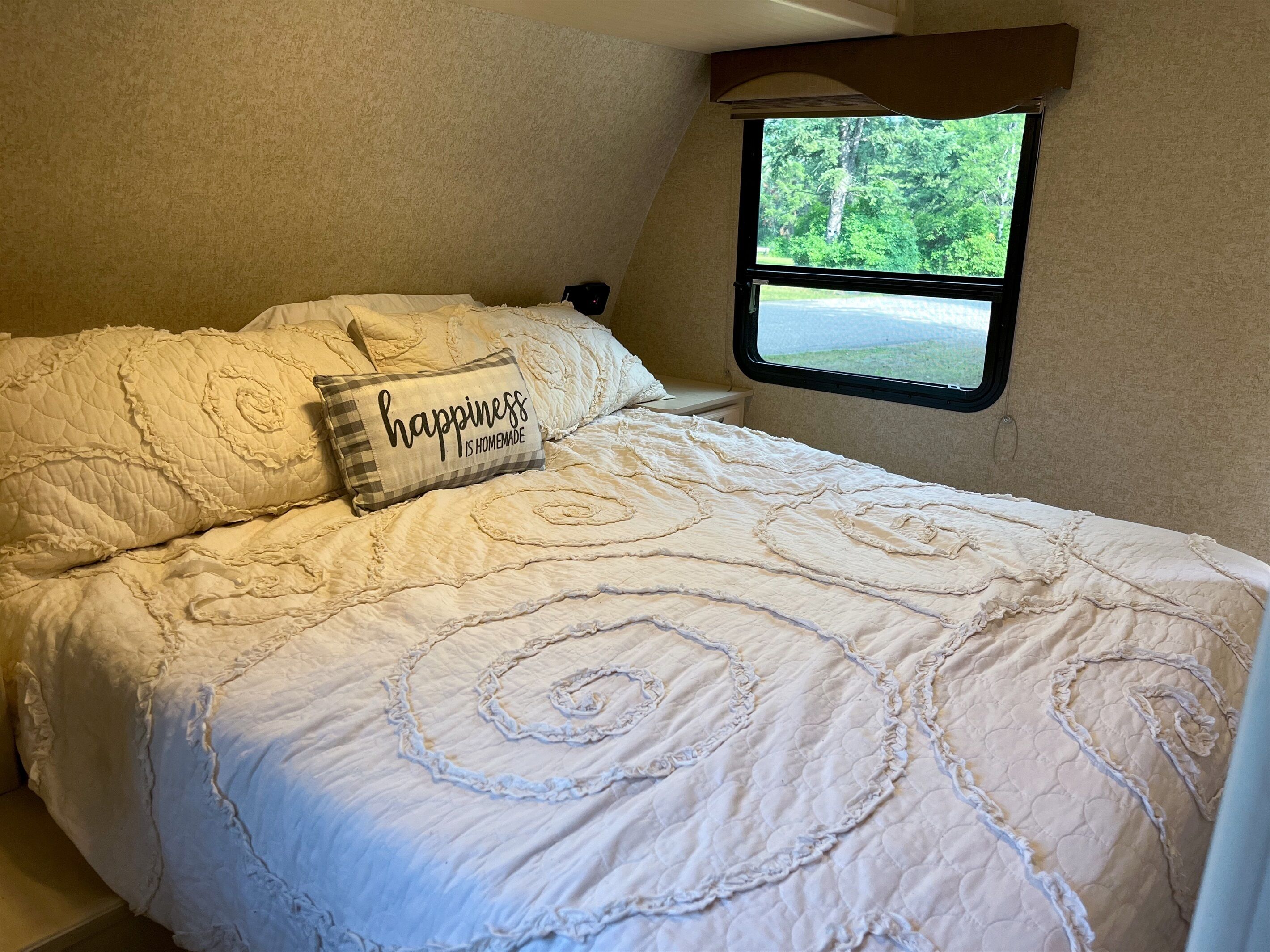 A bedroom with a bed, a pillow with the text "happiness is homemade," and a window with a view of trees.