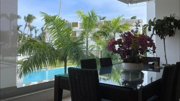 A modern room with a glass table and chairs, a large window with a view of a swimming pool and palm trees, and a ceiling fan. - Charming, luxurious 2 bedroom apt.  in amazing Las Terrenas with WiFi, AC.  (Las Terrenas)