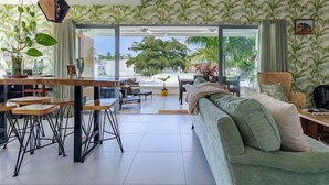 A modern living room with a green sofa, a dining area with stools, and large windows offering a view of palm trees. - Charming, luxurious 2 bedroom apt.  in amazing Las Terrenas with WiFi, AC.  (Las Terrenas)
