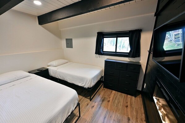 A compact bedroom with two single beds, a small dresser, a window with curtains, and a television.