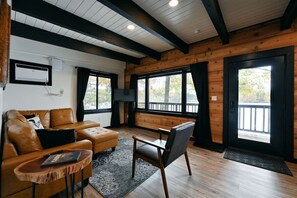 A cozy living room with a leather sofa, a wooden coffee table, and a chair. There are large windows with curtains, a ceiling fan, and an air conditioning unit on the wall.