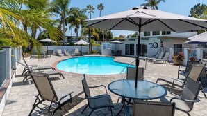 A swimming pool area with lounge chairs and umbrellas, surrounded by palm trees and a building with a clock. - Sandyland Beach Studio Carpinteria (Carpinteria)