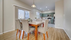 A dining area with a wooden table and chairs, a kitchen in the background, and a ceiling light fixture. - 4 bedroom fully Fenced 2+ Garage Pkg & Air Cond pet friendly  (Colorado Springs)