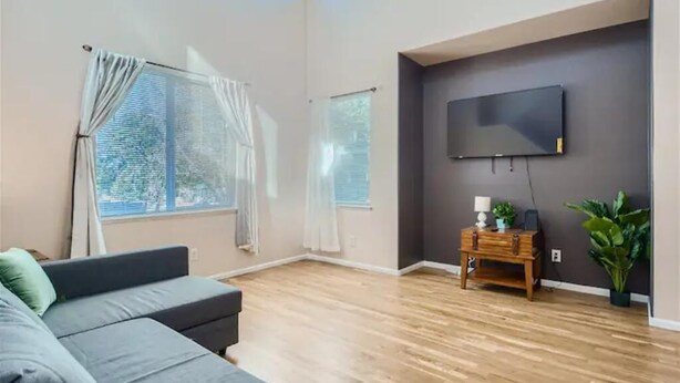 A modern living room with a grey sofa, wooden flooring, a mounted TV, and a small wooden side table with a lamp and plant. - 4 bedroom fully Fenced 2+ Garage Pkg & Air Cond pet friendly  (Colorado Springs)