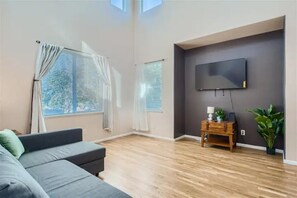 A modern living room with a grey sofa, wooden flooring, a mounted TV, and a small wooden side table with a lamp and plant.