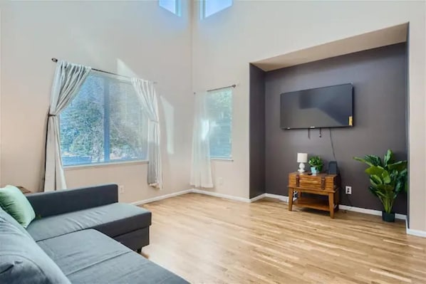 Un soggiorno moderno con un divano grigio, pavimento in legno, una TV a parete e un piccolo tavolino in legno con lampada e pianta.