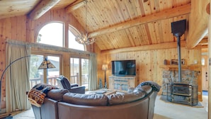 A cozy log cabin interior with a leather sofa, a television, a fireplace, and large windows. - Lake & Mtn Views: Spectacular Grand Lake Home (Grand Lake)