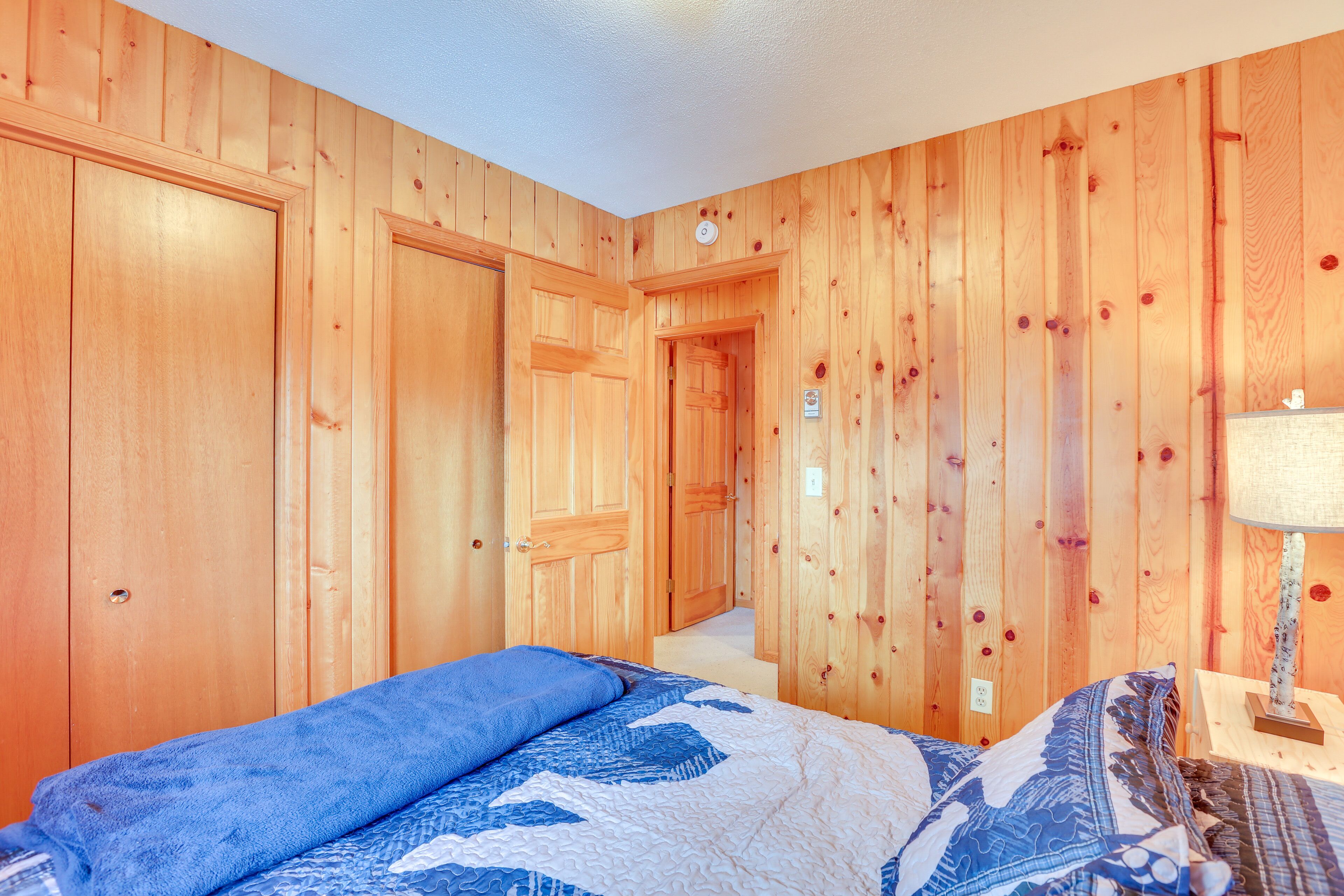 Un dormitorio con paredes de paneles de madera, una cama con ropa de cama azul y blanca, y una mesita de noche con una lámpara.