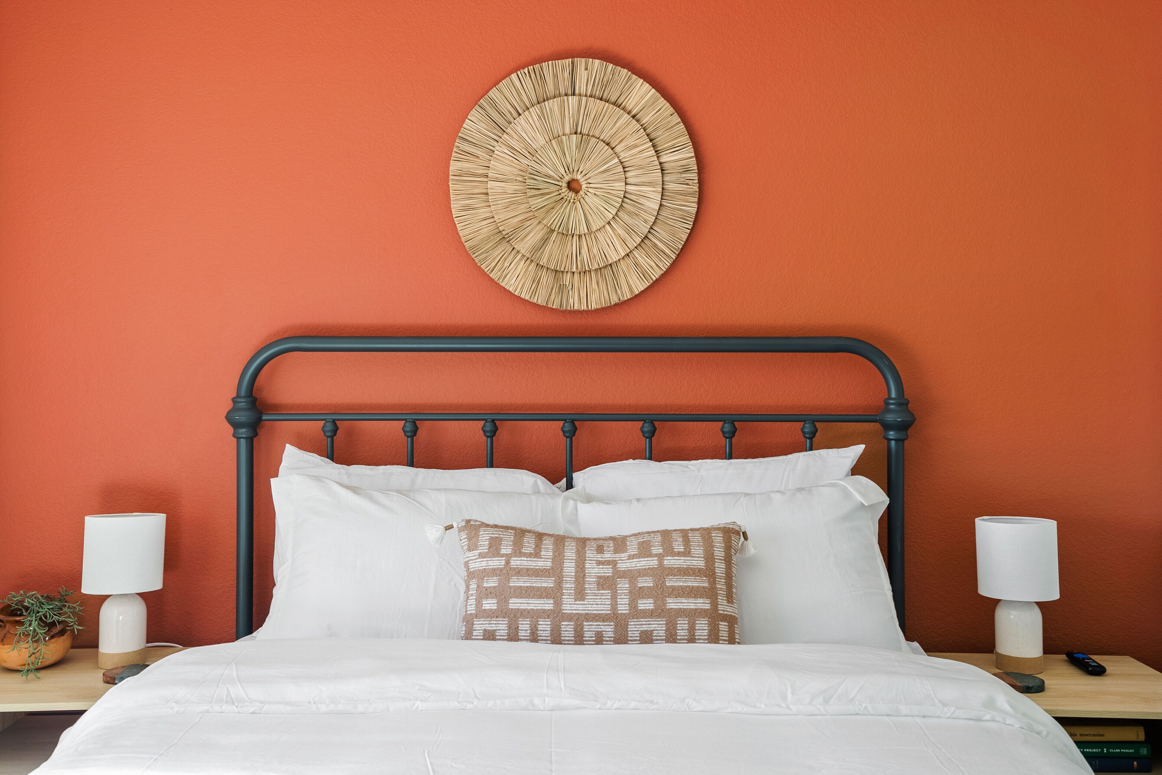 A neatly made bed with white linens and a patterned pillow, a round wall hanging, and two bedside lamps on a nightstand.
