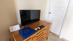 Un mueble de madera con un televisor de pantalla plana, un router blanco, una laptop azul y dos controles de videojuegos.