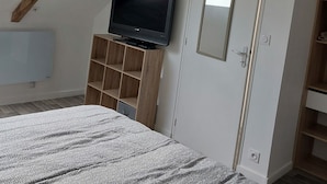Ein Schlafzimmer mit schrägverdeckter Decke, einem an einer Holzablage montierten Fernseher, einem Kleiderschrank und einem Bett mit gemusterten Bettbezügen.