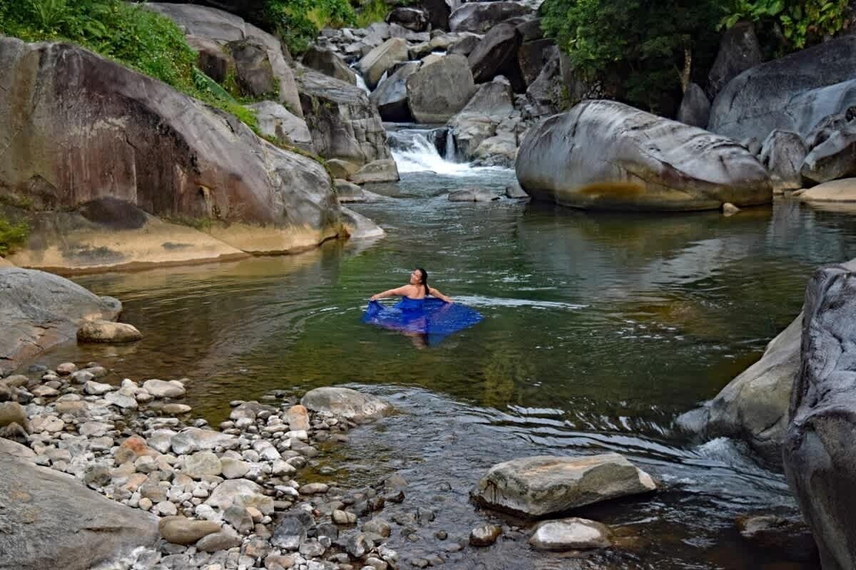 Uma pessoa flutua em uma jangada azul em um rio pedregoso, cercada por grandes pedras.