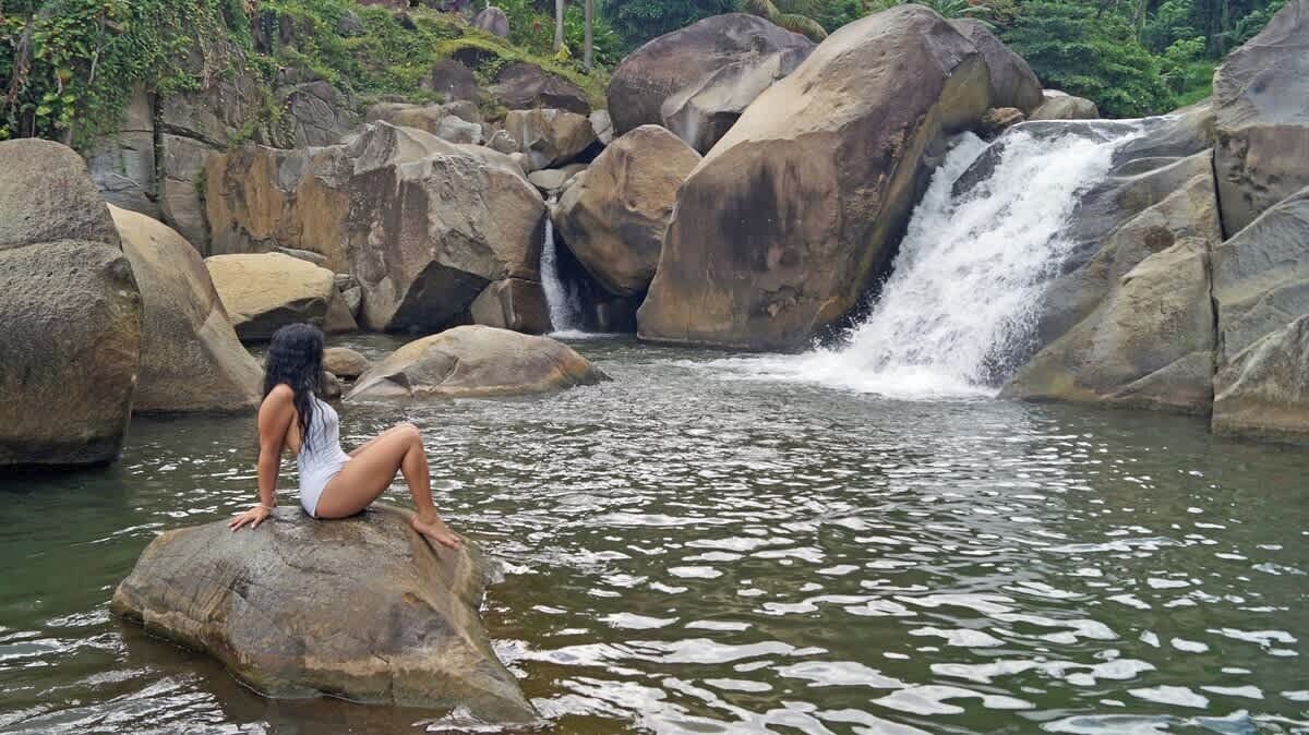 Eine Person sitzt auf einem Felsen neben einem Wasserfall in einer natürlichen Umgebung.