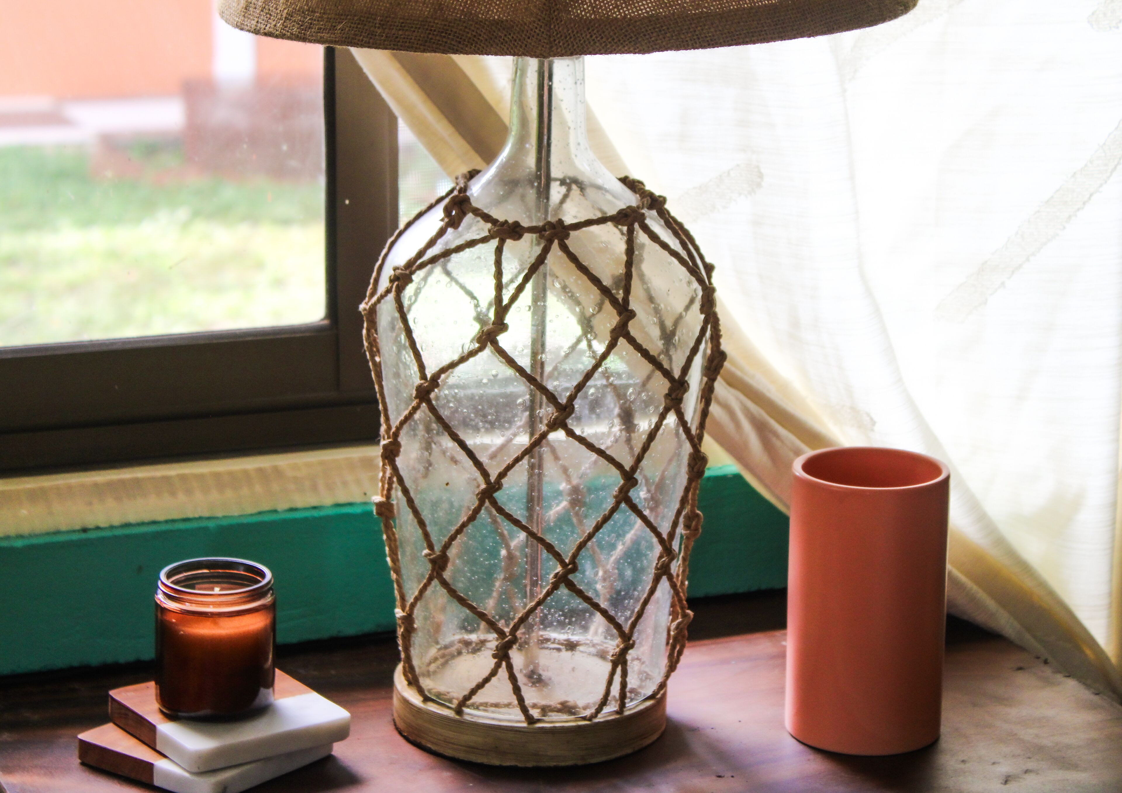 Um castiçal de vidro com padrão trançado, uma vela e uma chávena rosa num parapeito.