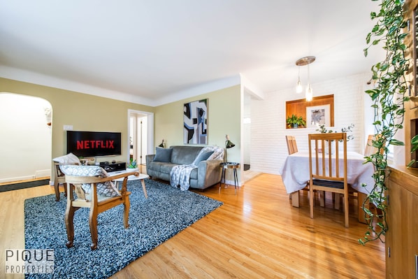 A living room with a TV displaying 'NETFLIX', a sofa, a coffee table, and a dining area with a table and chairs.