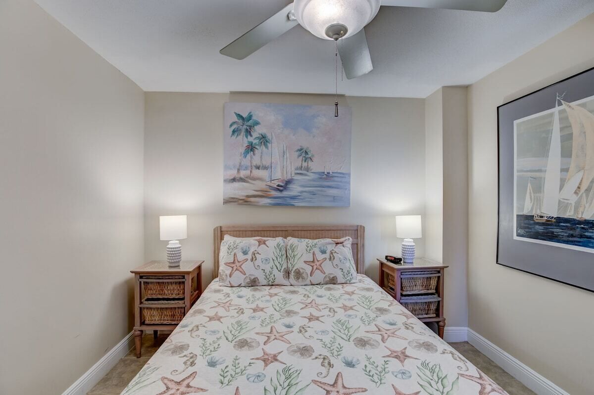 A bedroom with a bed, two bedside tables, a ceiling fan, and wall art depicting a beach scene.